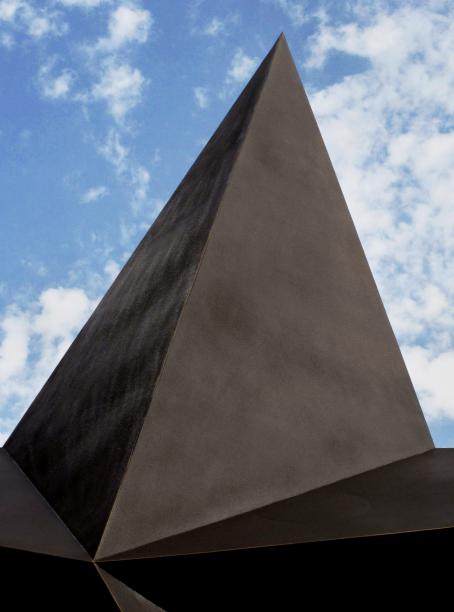 Ruskea, korkea, sileäpintainen pyramidin muotoinen rakennus, jonka alareunasta lähtee tasoja eri suuntiin. Rakennuksen takana näkyy sinistä taivasta ja valkoisia pilviä.