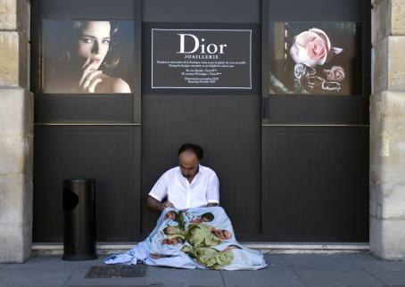 Mustahiuksinen mies istuu mustan seinän edessä ompelemassa lakanaa. Seinällä on mainoskuvia ja teksti "Dior".