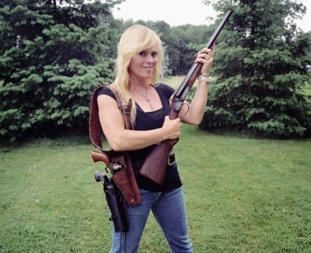 Vaaleahiuksinen nainen seisoo ulkona nurmikolla ja pitelee käsissään kivääriä. 