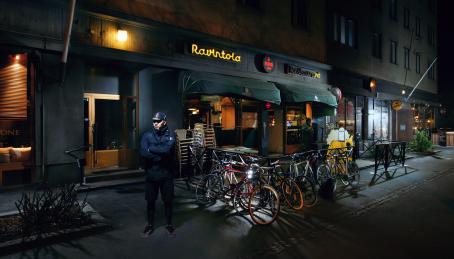 Hämärällä kadulla mies seisoo kädet puuskassa ravintolan edessä. Ravintolassa on terassi, ja terassin aitoihin on kiinnitetty polkupyöriä.