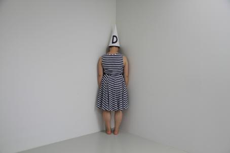 Valkoisen huoneen nurkassa seisoo selin raitamekkoinen nainen. Hänellä on päässään valkoinen kartionmuotoinen hattu, jossa on mustalla kirjain D.