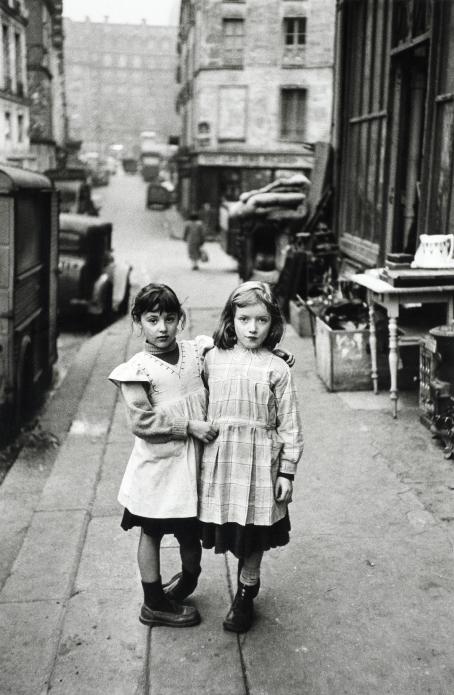 Mustavalkoisessa kuvassa kaksi pientä tyttöä kaupungin kadulla. Toinen pitää kättä toisen harteilla.