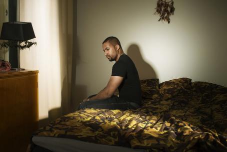 Tummaihoinen mies istuu sängyn reunalla. Sängylle tulee kaistale valoa. Seinällä sängyn yläpuolella roikkuu unisieppari. 