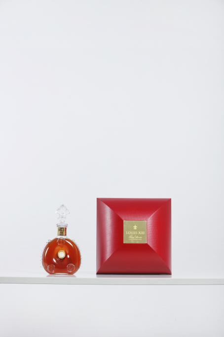 Valkoista taustaa vasten valkoisella tasolla on punainen laatikko ja sen vieressä koristeellinen läpinäkyvä pullo, jossa on punaista nestettä. Laatikon keskellä on kultainen laatta jossa lukee Louis XIII.