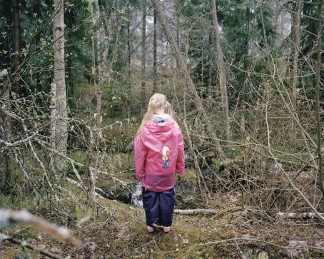 Metsässä pienen puron tai lammikon edessä seisoo selin tyttö, jolla on pitkissä, vaaleissa hiuksissa saparot ja pitkä vaaleanpunainen sadetakki.