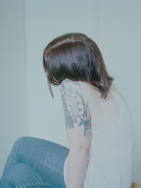 Nuori henkilö istuu sivuttain, hiukset peittävät kasvot. Hänellä on valkoinen toppi ja olkavarressa tatuointi.