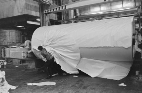 Mustavalkoisessa kuvassa on tehtaassa iso, monta metriä pitkä ja leveä paperirulla, josta työntekijä repii paperia irti.