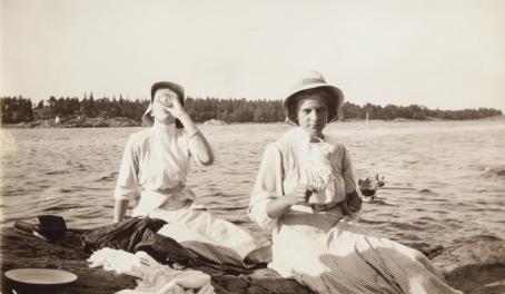 Mustavalkoisessa kuvassa kaksi naista istuvat rantakalliolla. Toinen on juuri juomassa lasista jotain, toisellakin on lasi kädessä. Heillä on päällään pitkät mekot ja hatut.