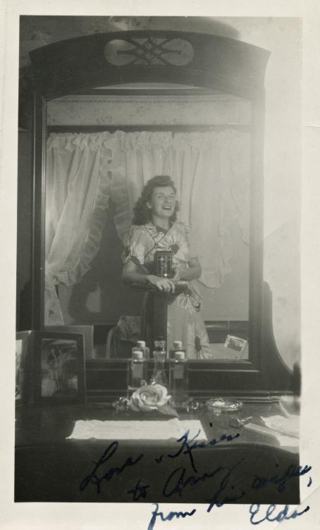 Mustavalkoinen kuva, jonka nainen on ottanut vanhanaikaisella kameralla kampauspöydän peilin kautta. Naisen mekossa on isojen kukkien kuvia ja hänellä on suu auki hymyssä. 
