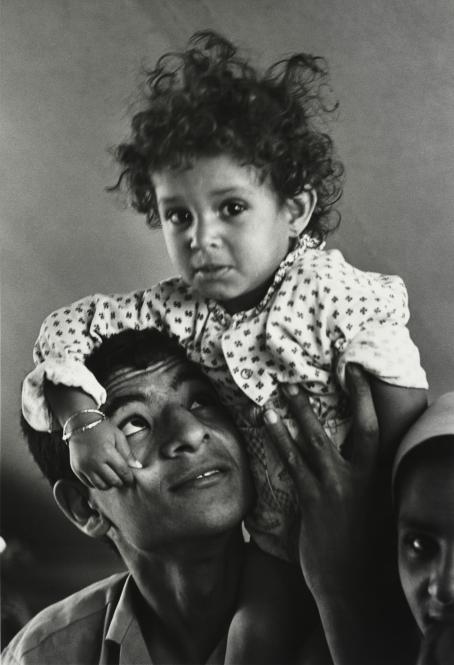 Syyria, 1982. Nuori mies sukulaislapsi olkapäällään Al-Sit Zeinab-nimisessä kaupungissa Damaskoksen lähellä. Mustavalkoisessa kuvassa miehen olkapäillä istuu pieni lapsi, joka puristaa miestä poskesta. Mies katsoo ylöspäin lapseen päin.