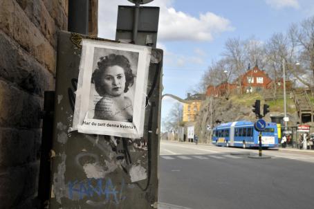 Seinän vieressä olevaan laatikkoon on teipattu paperi, jossa on mustavalkoinen naisen kuva ja sen alla teksti "Har du sett denna kvinna?". Taaempana näkyy bussipysäkki jolle bussi on pysähtynyt.
