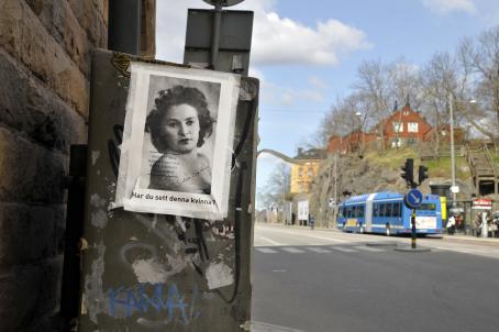 Seinän vieressä olevaan laatikkoon on teipattu paperi, jossa on mustavalkoinen naisen kuva ja sen alla teksti "Har du sett denna kvinna?". Taaempana näkyy bussipysäkki jolle bussi on pysähtynyt.