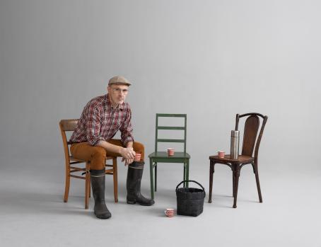 Mies istuu tuolissa kumisaappaat jalassa ja muki kädessään. Hänen vieressään on kaksi tuolia, joilla on mukit ja termospullo. Maassa on kori.