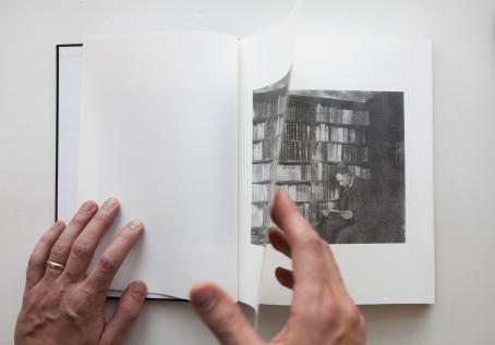 Kädet selaavat kirjaa. Kirjassa on mustavalkoinen kuva, jossa mies istuu ison kirjahyllyn edessä lukemassa kirjaa.