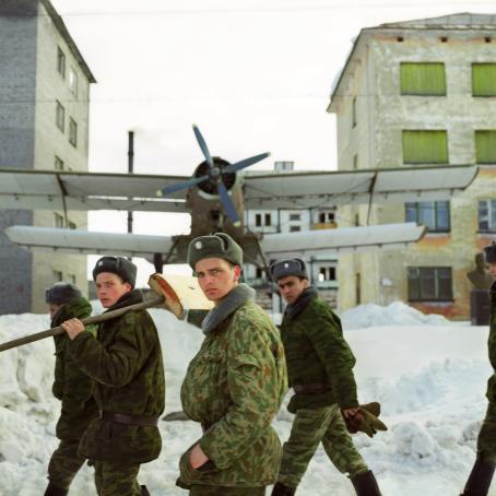 Armeijan asuihin pukeutuneita miehiä kävelee lumisella tiellä, yhden olalla on lumilapio. Heidän takanaan on potkurikone kerrostalojen välissä.