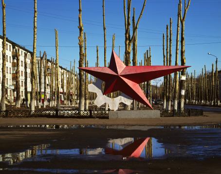 Monumentti, jossa on punainen tähti jonka sakarat ovat eripituisia, ja sen vieressä valkoinen hammasratas. Sen takana on lehdettomiä puita ja taaempana kerrostaloja.