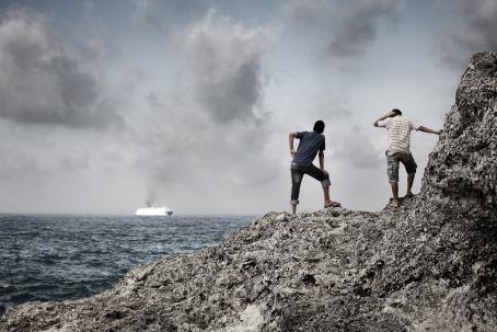 Kaksi ihmistä seisoo rantakalliolla selin kameraan. Toinen nojaa pystysuoraan kallioon ja toinen jalkoihinsa. Kauempana merellä menee risteilyalus.