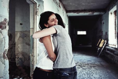 Kaksi ihmistä halaavat toisiaan. He ovat ränsistyneen näköisen rakennuksen käytävällä. Toinen heistä hymyilee leveästi, toisen kasvoja ei näy.