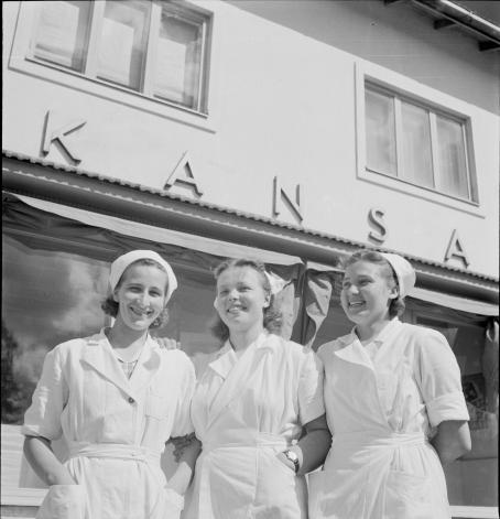 Kolme nuorta naista valkoisissa asuissa ja lakeissa. Takana olevan rakennuksen seinässä lukee "Kansa".
