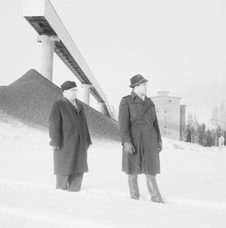 Kaksi miestä seisoo talvivaatteissa lumihangessa. Heidän takanaan näkyy kasa soraa tai jotain muuta ainetta ja pitkä, kapea pylväiden varassa oleva taso. 