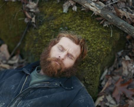 Mies jolla on oranssinruskeat hiukset ja parta makaa metsässä pää sammalen peittämällä kivellä. Hänellä on silmät kiinni ja päällä likainen sininen takki.