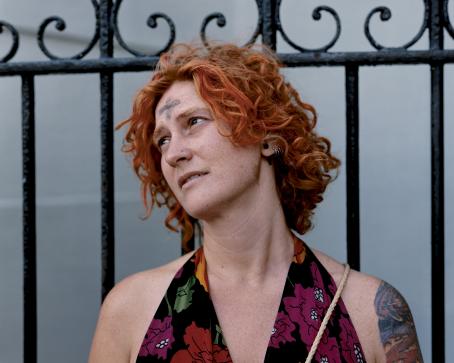 Nainen jolla on oranssit, kiharat hiukset, istuu nojaten metalliseen aitaan. Hänellä on otsassaan mustalla tehty risti ja olkavarressa tatuointi.