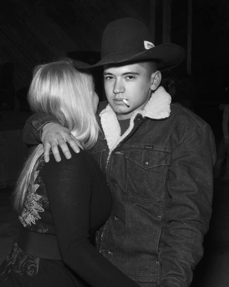 Mustavalkoisessa kuvassa mies karvavuorisessa farkkutakissa ja cowboy-hatussa, tupakka suussa. Hän halaa yhdellä kädellä vaaleahiuksista naista, naisen kasvot eivät näy kuvassa.