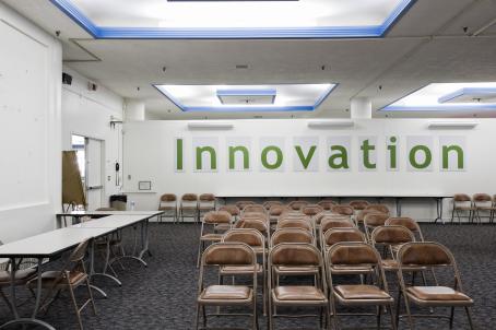 Huone, jossa on ruskeanahkaisia taittotuoleja monessa rivissä. Tilassa on sinisävyinen kokolattiamatto, sivuilla on pöytiä ja takaseinällä lukee Innovation.