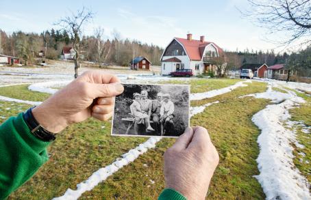 Omakotitalon pihapiiri, jossa on nurmikolla vähän lunta. Vanhat kädet pitävät mustavalkoista valokuvaa, jossa istuu kaksi lasta kahden aikuisen sylissä. 