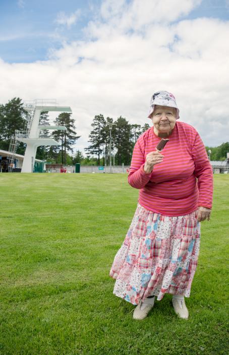 Vanha nainen seisoo nurmikolla vaaleanpunaraitaisessa paidassa ja vaaleanpunaisessa hameessa ja syö tikkujäätelöä. Taustalla näkyy uimahyppytorni.