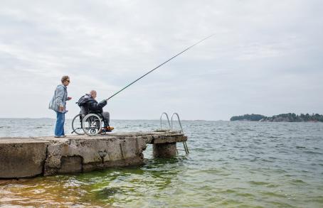 Vanha mies pyörätuolissa on onkimassa kivisellä laiturilla. Hänen vieressään on vanha nainen.