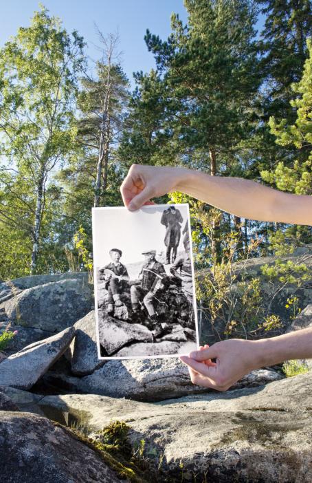Kallioita metsässä. Kädet pitelevät kallion edessä mustavalkoista valokuvaa, jossa pojat istuvat soittamassa samalla kalliolla kuin mikä taustalla on. Kuva on aseteltu niin, että kallion linjat täsmäävät.