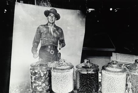 Mustavalkoisessa kuvassa isoja lasisia karkkipurkkeja rivissä. Niiden takana kuva cowboysta ase kädessä.