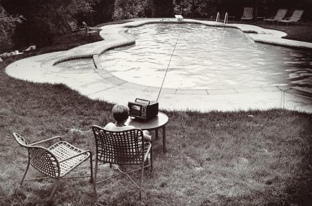 Mustavalkoisessa kuvassa uima-allas. Altaan vieressä nurmikolla istuu tuolissa lapsi kuuntelemassa pöydällä olevaa radiota.