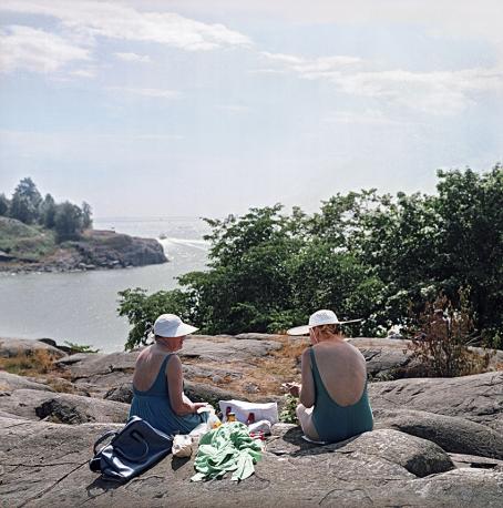 Kaksi naista istuu rantakallioilla. Toisella on uimapuku ja toisella mekko, heillä on valkoiset hatut päässä. Aurinko paistaa ja takana näkyy meri.