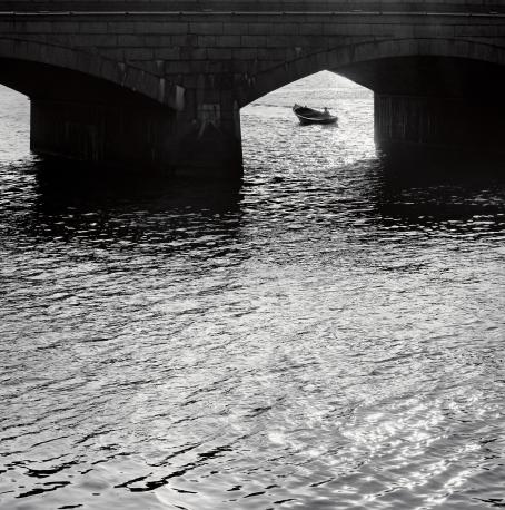 Mustavalkoisessa kuvassa etualalla on vettä, kuvan yläosassa näkyy osittain silta. Sillan alta kauempana näkyy vene.