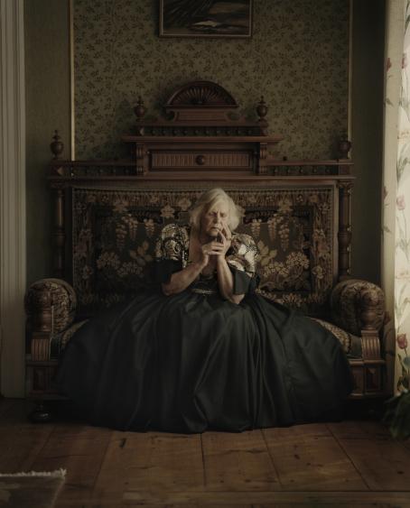 Vanha nainen istuu koristeellisella sohvalla, hänellä on päällään mekko jonka helma on lähes koko sohvan levyinen. Hän nojaa hieman päätään sormiinsa ja hänellä on silmät kiinni.