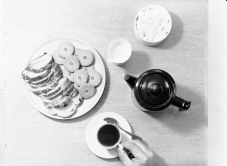 Mustavalkoisessa, ylhäältäpäin otetussa kuvassa on kahvipöytä. Käsi pitelee lautasen päällä olevaa kahvikuppia, lisäksi pöydällä on tarjoiluastiassa pullaa ja keksejä, sokeriastia, maito- tai kermakannu ja kahvipannu. 