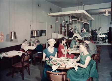 Näkymä kahvilasta sisältä. Etualalla pyöreässä pöydässä istuu kaksi nuorta naista mekot päällä. Toinen voitelee leipää. Taaempana näkyy lisää pöytiä ja kahvilan tiski. 