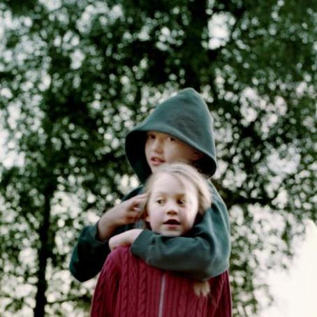 Lapsi jolla on vihreä huppari ja huppu päässä pitää kättään punapaitaisen lapsen ympärillä.