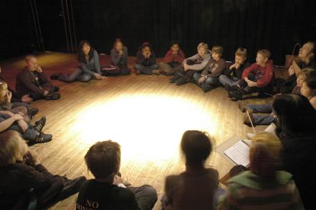 Lapsia istuu lattialla isossa ringissä. Heidän keskellään lattialla on iso valoympyrä, muuten huoneessa on melko hämärää. 