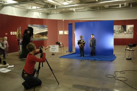 Ison tilan, ilmeisesti näyttelytilan, keskellä on pystyssä sininen kangas, jonka edessä seisoo kaksi lasta pukeutuneena roolivaatteisiin. Punapaitainen kuvaaja ottaa heistä kuvaa kolmijalalla olevalla kameralla. 
