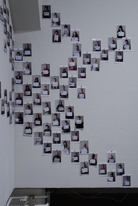 Seinälle epäsäännölliseen muotoon ripustettuja valokuvia. Kaikissa näyttäisi olevan henkilö pitelemässä paperia edessään. 
