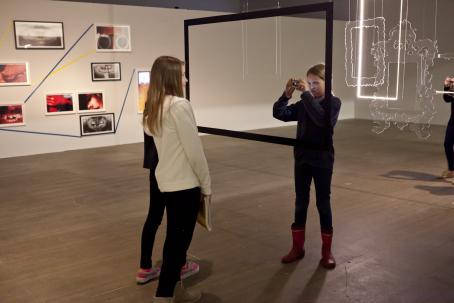 Näyttelytilassa nuori tyttö ottaa kuvaa toisesta tytöstä katosta roikkuvan, ison mustan kehyksen läpi. 