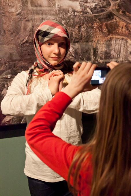 Nuori tyttö ottaa kuvaa toisesta tytöstä, joka seisoo taideteoksen edessä. Hänellä on ruudullinen huivi kiedottuna päänsä ympärille ja hän pitää käsiä ristissä rinnallaan.