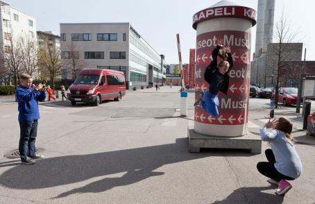 Tyttö on juuri hypännyt ilmaan. Hänen edessään on toinen tyttö maassa kyykyssä, he katsovat toisiinsa. Taaempana on sinipaitainen poika ottamassa kuvaa tilanteesta. 