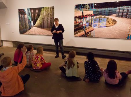 Lapsia istuu lattialla, yksi on nostanut kätensä ylös. Heidän edessään seisoo aikuinen, jonka takana seinällä on valokuvateoksia. 