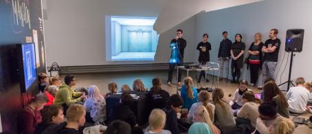 Darkroom Mansion -pelin lanseeraustilaisuus Suomen valokuvataiteen museossa lokakuussa 2017. Kuva: Virve Laustela / Suomen valokuvataiteen museo. Kuvan etualalla lattialla istuu lapsia. Taaempana seinälle on heijastettu kuva pelistä, ja mies puhuu mikrofoniin. Seinän vieressä seisoo aikuisia rivissä.