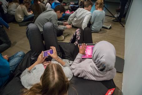 Takaapäin otettu kuva kahdesta tytöstä istumassa säkkituolilla. Toisella on puhelin ja toisella tabletti ja he molemmat pelaavat Darkroom Mansion-peliä. Taaempana lattialla istuu lisää lapsia. 