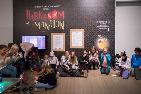 Darkroom Mansion -pelin lanseeraustilaisuus Suomen valokuvataiteen museossa lokakuussa 2017. Kuva: Virve Laustela / Suomen valokuvataiteen museo. Lattialla istuu lapsia, lähes kaikilla on kädessä puhelin tai tabletti jolla he pelaavat. Takana seinällä on tumma tapetti ja siinä lukee Darkroom Mansion.